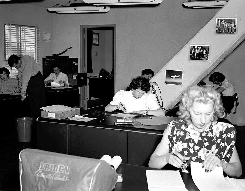 black and white photo of a NACA computer room from the 40s, showing women working at desks with a Friden mechanical calculator in the foreground, papers scattered around and photos of planes on the wall